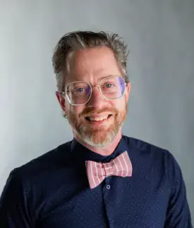 James Sansbury smiling, sporting a pink bowtie and nerdy glasses.