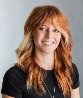 A white woman with long red hair wearing a black shirt top in front of a gray background