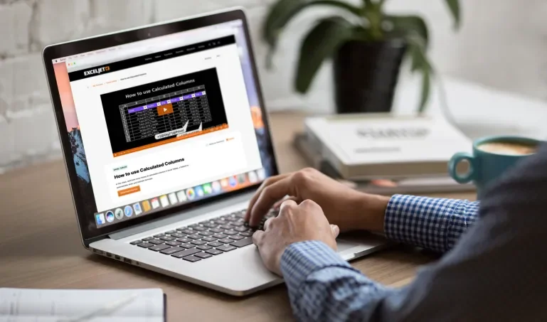 Image of hands resting on an open laptop on a desk with the Exceljet website on the screen, small green plant, some books, papers, and a blue coffee mug on the desk near the laptop.