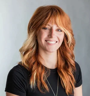 A white woman with long red hair wearing a black shirt top in front of a gray background