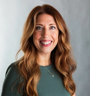 A white woman with red hair, standing in front of a gray background.