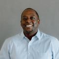 Albert Hughes, an African American man wearing a blue button down oxford shirt in front of a gray backdrop.