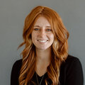 Photo of Alycia Jones, white women with long red hair wearing a black v-neck top in front of a gray background.