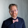 A photo of James Sansbury smiling, sporting a pink bowtie and nerdy glasses.