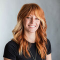 A white woman with long red hair wearing a black shirt top in front of a gray background